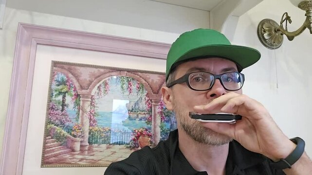 MOSCOW - NOV 15, 2023: Man (MR) wearing a green hat and glasses is holding a harmonica mouthorgan to his mouth and play music