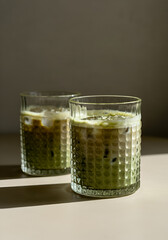 Two textured glass cups filled with iced matcha latte, resting on a light matte surface in a clean indoor environment. 