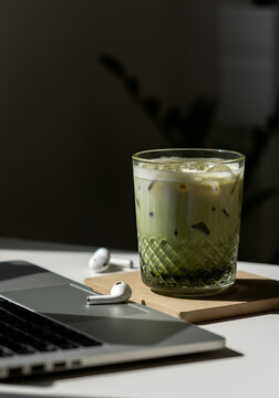 A textured glass of iced matcha latte, a pair of white wireless earbuds with case, and a black smartphone arranged diagonally on a clean white surface. 