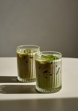 Two textured glass cups filled with iced matcha latte, resting on a light matte surface in a clean indoor environment. 