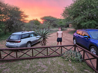 Man stands in front of a fence with a car behind him. The car is blue and is parked in front of a...