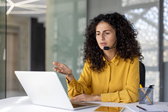 A woman with curly hair wearing a headset and yellow shirt, looks at a laptop while gesturing, indicating a business meeting or customer service interaction.