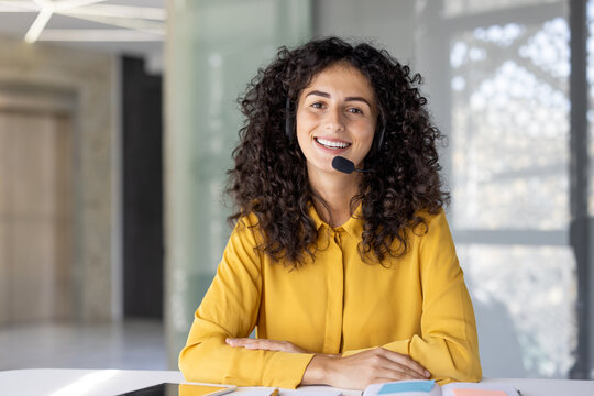 A friendly customer service representative with curly hair smiles while wearing a headset in a bright office setting.