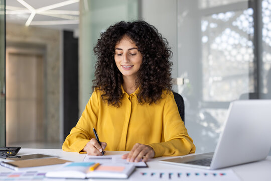 A smiling businesswoman in a yellow shirt works at her desk, writing on paper with a pen.