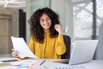 A happy woman with curly hair celebrates a success at her desk, holding documents and working on...