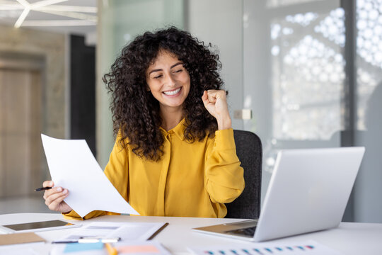A happy woman with curly hair celebrates a success at her desk, holding documents and working on her laptop.