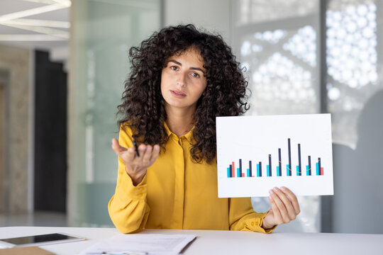A businesswoman presents a graph, gesturing with a pen while looking at the camera in an office setting.