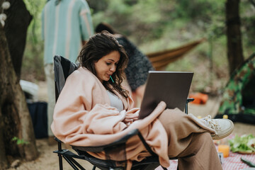 A young woman sits in a tranquil forest while using her laptop. Wrapped in a blanket, she enjoys...