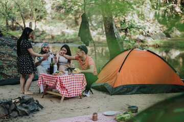Group of friends camping near a calm riverbank, sharing drinks and food at a picnic table while...