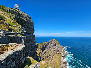 Lighthouse is on top of a cliff overlooking the ocean. The sky is clear and blue. The ocean is calm...