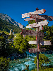 Rustic Wooden Signpost in Traditional Swiss Village of Splugen: Charming Mountain Scenery with Alpine Houses, Church Tower and Flowing River Beneath Majestic Peaks