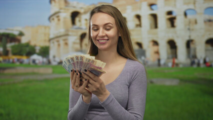 Woman holds polish zloty banknotes in front of the roman coliseum, smiling with a joyful expression, suggesting travel and financial success in italy.
