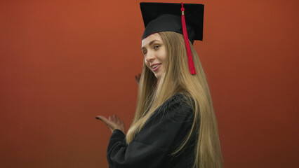 Woman graduate in cap and gown pointing to side over red background showcasing academic achievement...