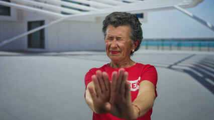 Senior woman with grey hair in red shirt gesturing stop at seaside promenade during daylight,...