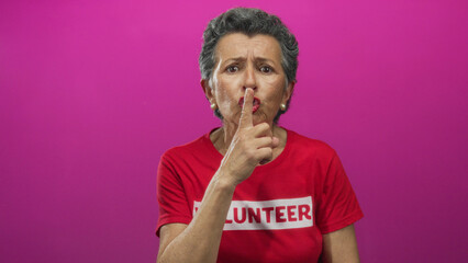 Senior woman with grey hair wearing a volunteer shirt gestures for silence against a vibrant pink...