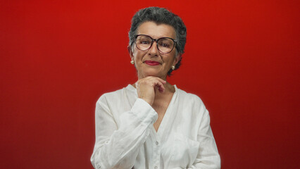 Obraz premium Senior woman with grey hair and glasses in a white shirt posing confidently against an orange background, exuding elegance and wisdom with a thoughtful expression.
