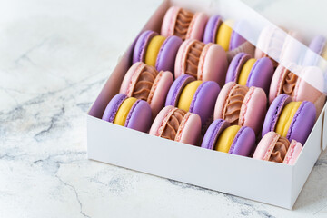Close up of multicolored macaroons in the cardboard box on the white marble background. Assortment of pink and violet macaroon with yellow chocolate filling. Famous French dessert in a gift box