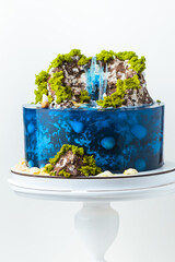 Beautiful birthday cake in the shape of an island decorated with chocolate edible mountains, greenery with waterfall. Tropical island cake surrounded with blue jelly ocean water