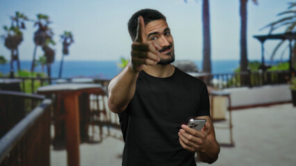 Young hispanic man pointing at camera with smartphone outdoors by the sea under palm trees, exuding a casual and approachable vibe on a sunny day.