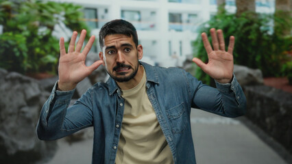 Naklejka premium Young man with beard standing outdoors in urban area expressing surprise against a backdrop of greenery and buildings in daylight, wearing casual denim and beige clothing.