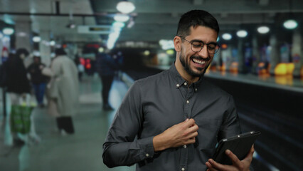 Young man using a tablet in a busy indoor train station, smiling, wearing glasses, with people in...