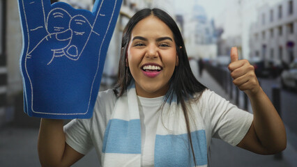 Young woman enthusiastically supports team outdoors with a foam finger and thumbs up, showcasing...