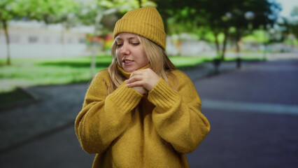 Woman shivering in yellow sweater and knit hat in outdoor park setting, expressing cold, with trees...