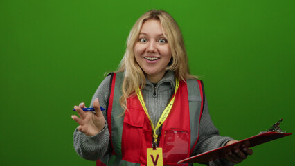 Young woman volunteer with blonde hair in red vest holding clipboard and pen over green background...