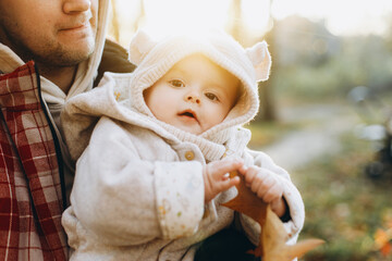 Happy cute baby son holding autumn leaf in fathers hands in soft evening light in autumn woods. Fatherhood. Stylish man with adorable child in cozy warm sweaters hugging in fall park