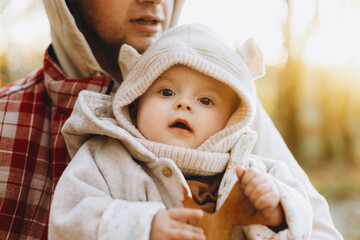 Happy cute baby son holding autumn leaf in fathers hands in soft evening light in autumn woods. Fatherhood. Stylish man with adorable child in cozy warm sweaters hugging in fall park