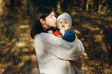 Happy mother hugging and kissing cute baby son in autumn woods. Motherhood. Stylish woman with adorable child in cozy warm sweaters relaxing in fall park