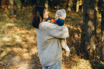 Happy mother hugging cute baby son in autumn woods. Motherhood. Stylish woman with adorable child in cozy warm sweaters embracing and relaxing in fall park