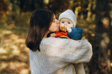 Happy mother hugging and kissing cute baby son in autumn woods. Motherhood. Stylish woman with adorable child in cozy warm sweaters relaxing in fall park