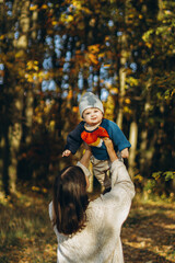 Happy mother playing with cute baby son in autumn woods. Motherhood. Stylish woman with adorable child in cozy warm sweaters having fun and relaxing in fall park