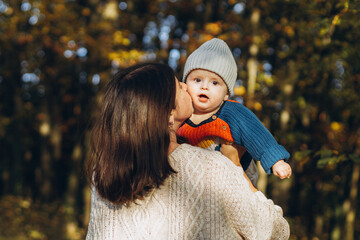 Happy mother hugging and kissing cute baby son in autumn woods. Motherhood. Stylish woman with adorable child in cozy warm sweaters embracing and relaxing in fall park