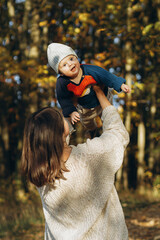 Happy mother playing with cute baby son in autumn woods. Motherhood. Stylish woman with adorable child in cozy warm sweaters having fun and relaxing in fall park