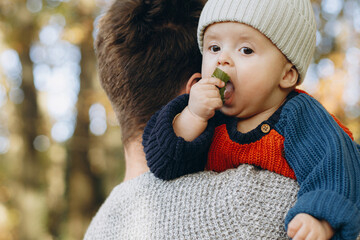 Cute baby son with funny emotions holding leaves close up in father hands  in autumn woods. Fatherhood. Stylish man with adorable child in cozy warm sweaters relaxing in fall park