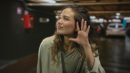 Young woman presses hand to ear listening gesture in underground parking building next to parked...