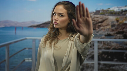 Woman with serious expression extends palm for stop on seaside promenade; defiance refusal boundary.