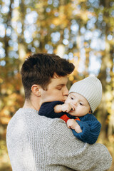 Happy father hugging and kissing cute baby son in autumn woods. Fatherhood. Stylish man with adorable child in cozy warm sweaters embracing and relaxing in fall park