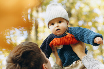 Cute baby son with funny emotions in father hands close up in autumn woods. Fatherhood. Stylish man with adorable child in cozy warm sweaters having fun and relaxing in fall park