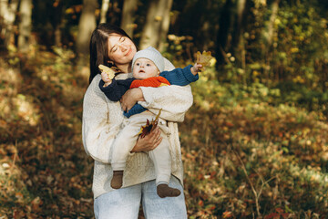 Happy mother hugging cute baby son in autumn woods. Motherhood. Stylish woman with adorable child in cozy warm sweaters embracing and relaxing in fall park