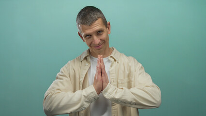 Man smiling with hands in prayer gesture against blue wall wearing casual shirt isolated in studio setting signifying peace and gratitude