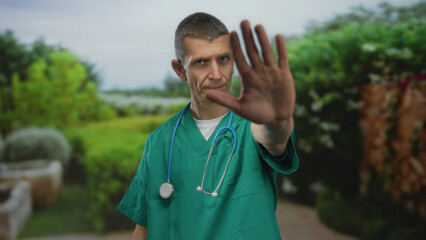 Doctor in green uniform with a stethoscope gestures stop in a park, surrounded by lush greenery,...