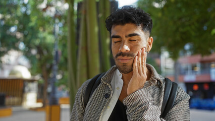 Young man with beard holding cheek in pain at a city park, showing toothache in an urban outdoor...