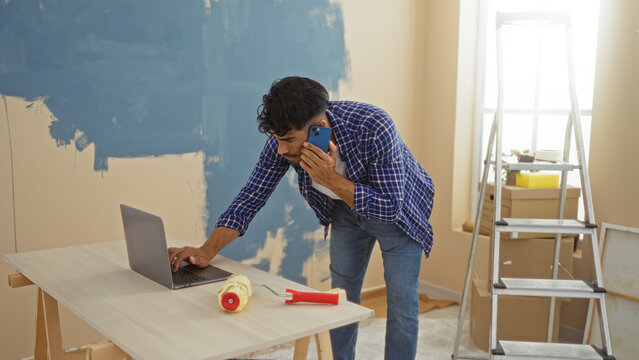 Young man with beard working on laptop during home renovation in living room, surrounded by paint, ladder, and boxes, multitasking with phone and computer. - Powered by Adobe