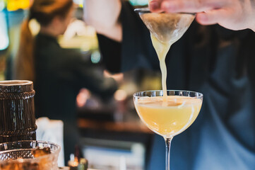 Bartender pouring a yellow cocktail through a strainer into a glass in a stylish bar setting. Concept of mixology, nightlife, and professional drink preparation.