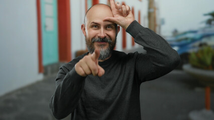 Bearded man gesturing loser sign with hand on forehead in urban street setting, showcasing humor...