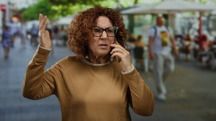 Woman gesturing emotionally while talking on a phone in a bustling urban street setting, showing...