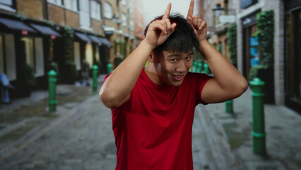 Obraz premium Young man in red shirt making a horns gesture while standing in a lively urban street setting, surrounded by colorful buildings and a cheerful atmosphere.